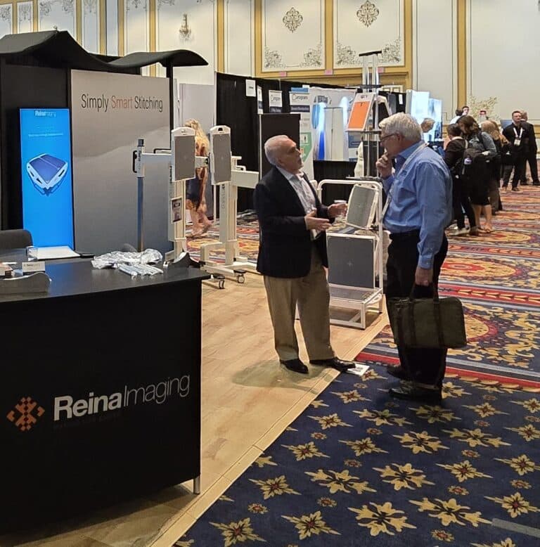 Trade show scene where two attendees talk beside a Reina Imaging booth showcasing medical imaging equipment and displays in a busy convention hall.