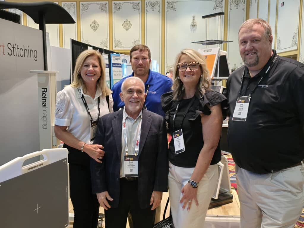 Group photo of five people standing together at a Reina Imaging trade show booth, surrounded by medical imaging equipment and displays inside a convention hall.