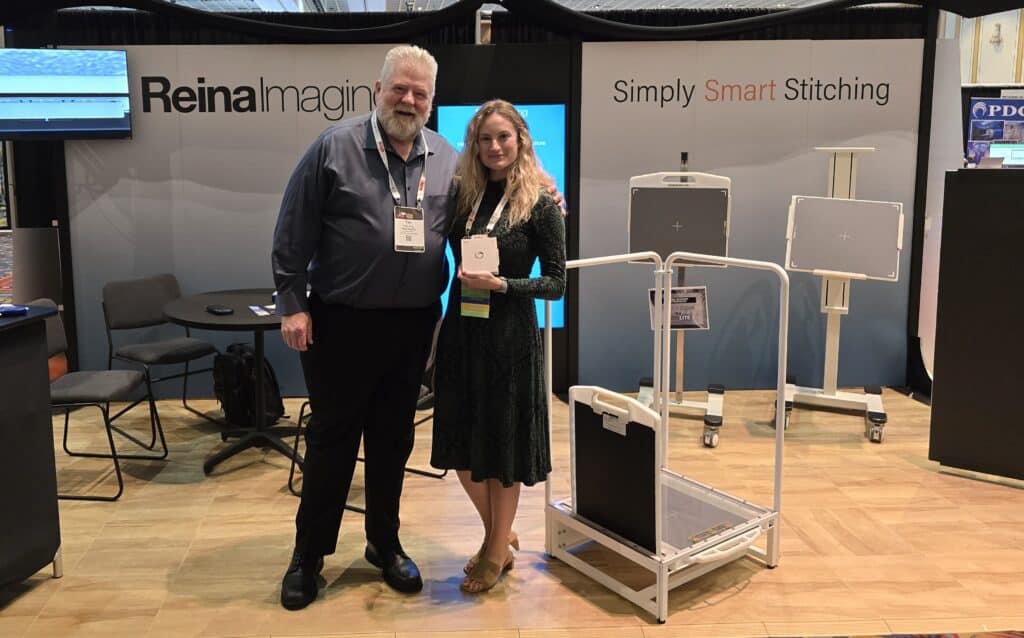 Two people pose together at a Rein Imaging trade show booth, standing near medical imaging equipment and branded displays inside a convention hall.