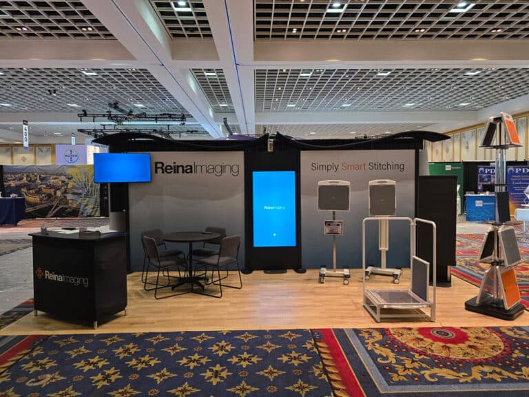 Trade show booth displaying Reina Imaging branding, digital screens, medical imaging equipment, and a small seating area inside a convention hall.