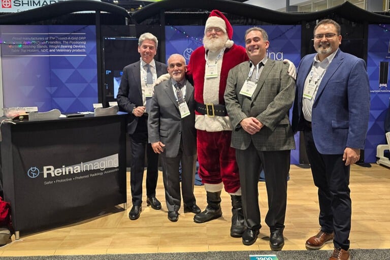 Group of adults posing together at a trade show booth with branded signage and displays, including one person dressed in a Santa Claus costume in an exhibition hall.