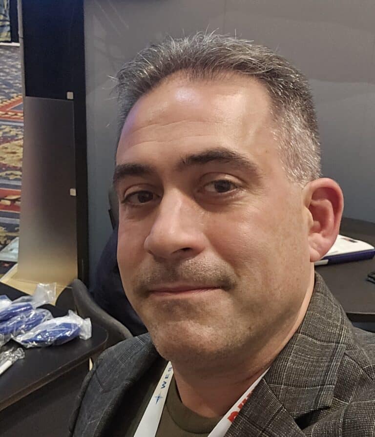 Close-up photo of a person wearing a jacket and lanyard, standing indoors near a trade show booth with materials and displays visible in the background.